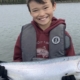 Boy wearing a life jacket on a boat holding a large silver fish, smiling at the camera near a lake edge.