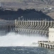 Dam spillway with multiple sluice gates releasing water, creating mist near a concrete promenade and rocky hills in the background