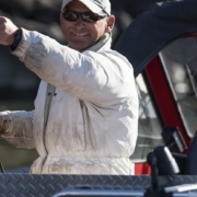 Man wearing a light jacket and sunglasses on a boat, proudly holding up a small fish he caught.