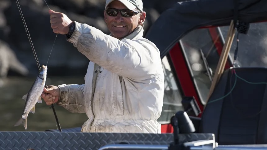 Man wearing a light jacket and sunglasses on a boat, proudly holding up a small fish he caught.