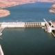 Aerial view of a concrete dam across a wide desert river, with spillways and a gated intake structure extending into the water.