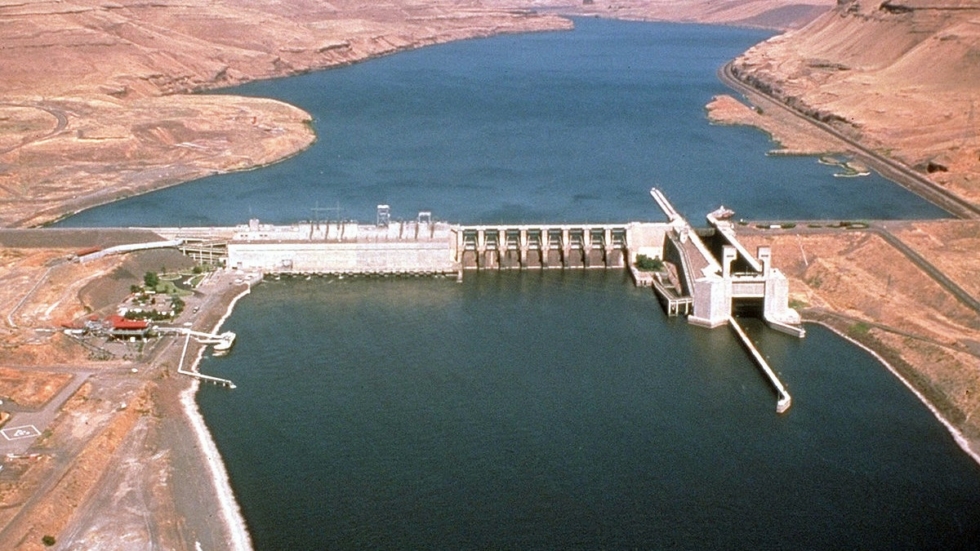 Aerial view of a concrete dam across a wide desert river, with spillways and a gated intake structure extending into the water.
