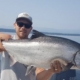 Person wearing a cap and sunglasses on a boat holds a large, silver fish horizontally across their chest, with a calm lake in the background.