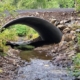 Concrete arch culvert under a railroad track with a shallow stream flowing through among rocks and green foliage on the banks.