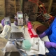 Two researchers in PPE work at a cluttered lab bench with tubes, pipettes, and a small centrifuge.