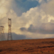 Transmission towers and a wind turbine on a brown hill under a dramatic cloudy sky, with power lines crossing the scene.