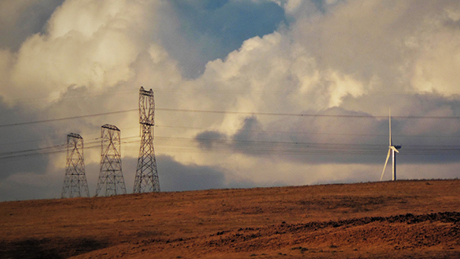 Transmission towers and a wind turbine on a brown hill under a dramatic cloudy sky, with power lines crossing the scene.