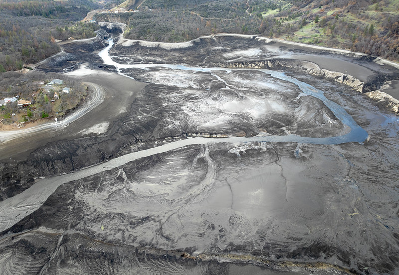 Aerial view of a fresh lava field with dark cooled lava, braided channels, and a small settlement on the left hillside. The area shows a pale crust and a blue water-like channel weaving through the flows.