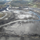 Aerial view of a fresh lava field with dark cooled lava, braided channels, and a small settlement on the left hillside. The area shows a pale crust and a blue water-like channel weaving through the flows.