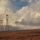 Wide view of a grassy hill with power pylons on the left and a wind turbine on the right beneath a cloudy sky.