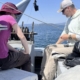 Two people on a boat handling fishing gear with reels, blue water and hills in the background.