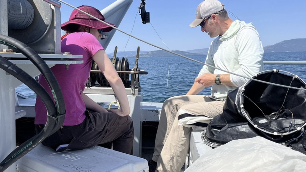 Two people on a boat handling fishing gear with reels, blue water and hills in the background.