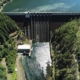 Concrete dam with spill gates releasing water, surrounded by forested hills and a small building nearby.