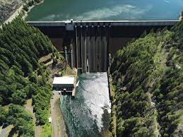 Concrete dam with spill gates releasing water, surrounded by forested hills and a small building nearby.