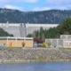Hydroelectric dam with buildings along a rocky riverbank, forested hills in the background.