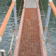 Industrial metal grate walkway with orange handrails spanning a green water channel, extending toward a distant platform.
