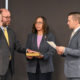 Man raises right hand to take an oath, with a woman holding a yellowed book and a man presenting a document in a formal swearing-in ceremony, flags in the background.
