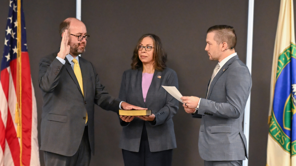 Man raises right hand to take an oath, with a woman holding a yellowed book and a man presenting a document in a formal swearing-in ceremony, flags in the background.