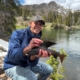Smiling man in a blue jacket kneels by a lake, holding a large fish with a rocky, forested mountainside in the background.