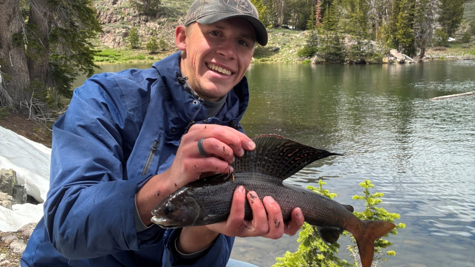 Smiling man in a blue jacket kneels by a lake, holding a large fish with a rocky, forested mountainside in the background.